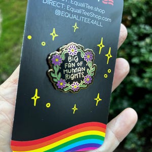 Pu&ograve; includere: Spilla in smalto nero con la scritta "Big Fan of Human Rights" circondata da una ghirlanda floreale con fiori viola. La spilla &egrave; su una carta nera con un arcobaleno e la parola "EQUALITY" in alto.