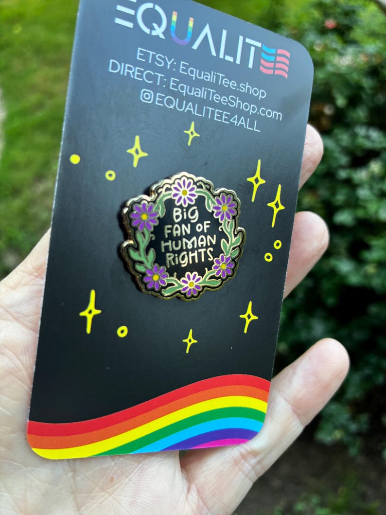 Pu&ograve; includere: Spilla in smalto nero con la scritta "Big Fan of Human Rights" circondata da una ghirlanda floreale con fiori viola. La spilla &egrave; su una carta nera con un arcobaleno e la parola "EQUALITY" in alto.