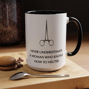 May include: White ceramic mug with a black interior and handle. The mug features a black illustration of surgical scissors and the text: "NEVER UNDERESTIMATE A WOMAN WHO KNOWS HOW TO NEUTER."