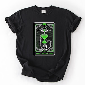 May include: Black t-shirt featuring a tarot card design with a green and white color scheme. The card depicts a skeleton holding a chalice, with an eye above. The text "THE COLLECTOR" is at the bottom.
