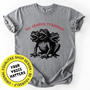 May include: A gray t-shirt featuring a black illustration of a three-headed frog with the text "SIC SEMPER TYRANNIS" in red above it. A yellow and white circular logo is in the lower left corner with the text "YOUR VOICE MATTERS".