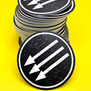 May include: A stack of round black and white stickers with three white arrows pointing down and to the left. The stickers are on a bright yellow surface.