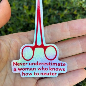 May include: A holographic sticker in the shape of surgical scissors with red accents. The sticker has the text "Never underestimate a woman who knows how to neuter" in red letters.