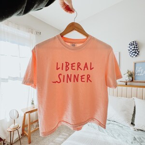 May include: A peach-colored t-shirt with the words "LIBERAL SINNER" printed in red. The shirt is hanging on a wooden hanger. The text is in a bold, sans-serif font.