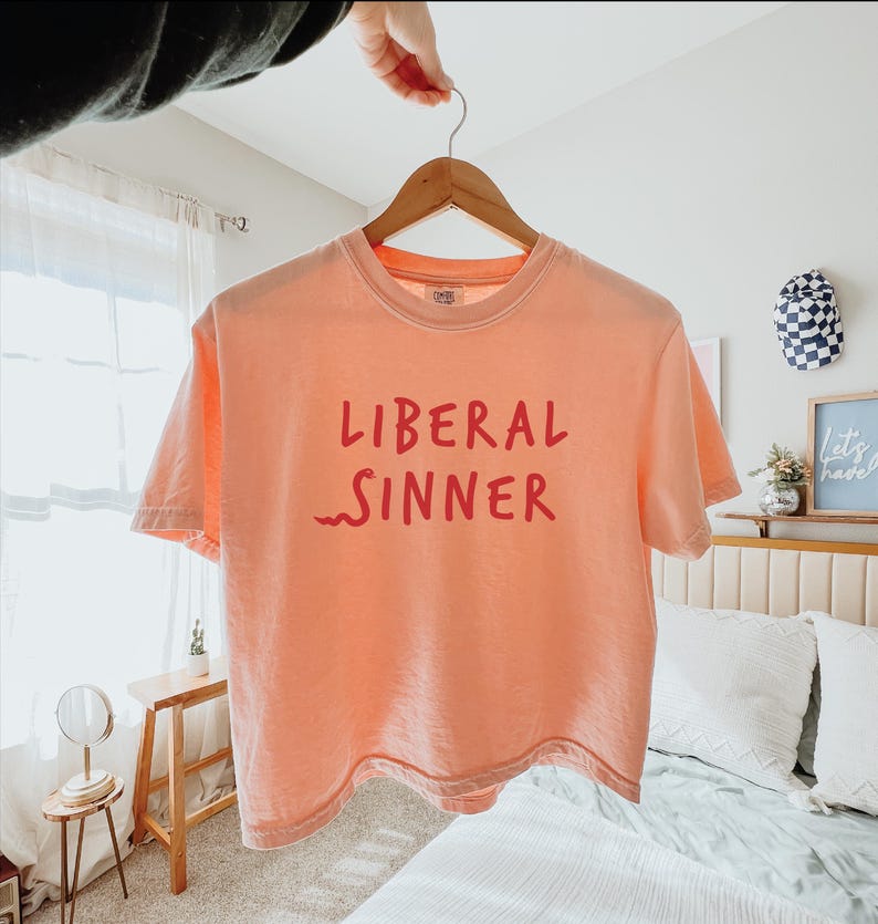 May include: A peach-colored t-shirt with the words "LIBERAL SINNER" printed in red. The shirt is hanging on a wooden hanger. The text is in a bold, sans-serif font.