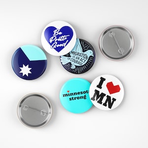 May include: A collection of round pinback buttons with various designs and text. One button has a blue heart with "Be Pretty Good" text. Another says "Migration is an Act of Courage". Other buttons say "Minnesota strong" and "I ❤️ MN".