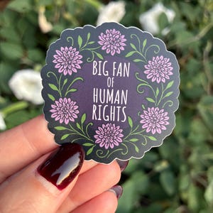 May include: A sticker with a dark purple background and the text "BIG FAN OF HUMAN RIGHTS" in white. The sticker is surrounded by pink flowers and green vines. The sticker is held by a person.