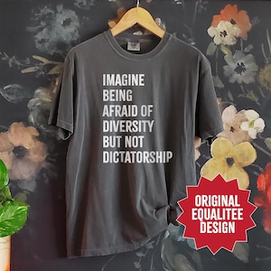May include: A gray t-shirt with the text "IMAGINE BEING AFRAID OF DIVERSITY BUT NOT DICTATORSHIP" in white. The shirt is hanging on a wooden hanger. A red starburst graphic reads "ORIGINAL EQUALITEE DESIGN".