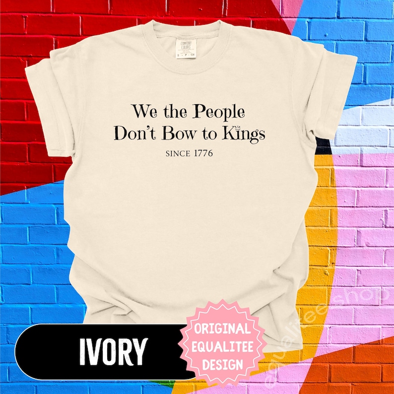 K&ouml;nnte beinhalten: Elfenbeinfarbenes T-Shirt mit dem schwarzen Text "We the People Don't Bow to Kings SINCE 1776". Das Shirt hat einen Rundhalsausschnitt und kurze &Auml;rmel. Ein rosa-wei&szlig;er Aufkleber mit "ORIGINAL EQUALITEE DESIGN" befindet sich unten rechts.