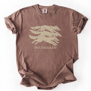 May include: A brown t-shirt featuring a graphic of three running foxes in a light tan color. Below the foxes is the Spanish phrase "&iexcl;NO PASAR&Aacute;N!" in a matching light tan color. The shirt has a crew neck and short sleeves.