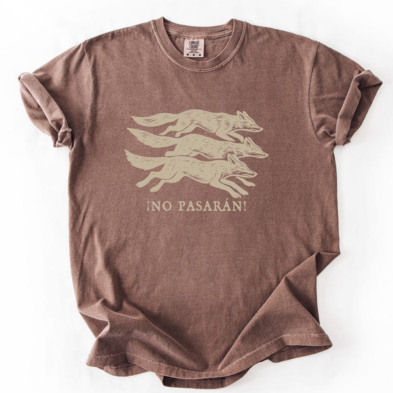 May include: A brown t-shirt featuring a graphic of three running foxes in a light tan color. Below the foxes is the Spanish phrase "&iexcl;NO PASAR&Aacute;N!" in a matching light tan color. The shirt has a crew neck and short sleeves.