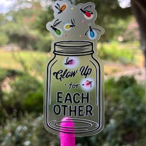 May include: A clear sticker in the shape of a mason jar with the words "Glow Up for Each Other." The sticker has a pink pen attached to the bottom. The top of the sticker has several colorful fireflies.