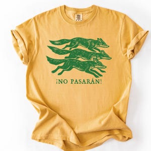May include: Mustard yellow t-shirt featuring a green graphic of three running foxes. Below the foxes, the text "&iexcl;NO PASAR&Aacute;N!" is printed in green. The shirt has a crew neck and short sleeves.