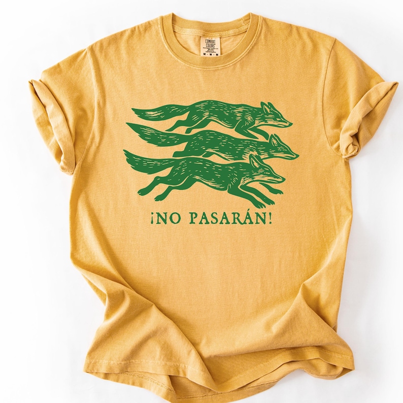 May include: Mustard yellow t-shirt featuring a green graphic of three running foxes. Below the foxes, the text "&iexcl;NO PASAR&Aacute;N!" is printed in green. The shirt has a crew neck and short sleeves.