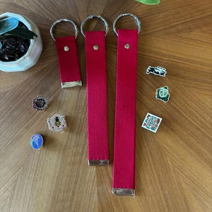 May include: Three red fabric wristlet keychains of varying lengths, each featuring a silver-tone metal ring and a silver-tone metal clamp at the base. Several small enamel pins are scattered nearby, including designs of a bee, a frog, and text.