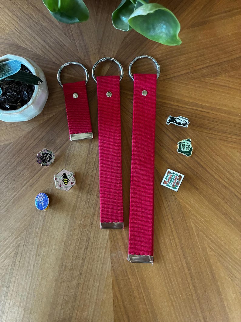 May include: Three red fabric wristlet keychains of varying lengths, each featuring a silver-tone metal ring and a silver-tone metal clamp at the base. Several small enamel pins are scattered nearby, including designs of a bee, a frog, and text.