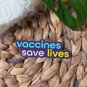 May include: A sticker with the text "vaccines save lives" in blue, pink, and yellow, set against a dark blue background. The sticker is placed on a woven, brown surface, with a green leaf and white fluffy material in the background.