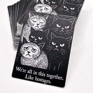 May include: A deck of black cards featuring illustrations of grumpy cats. The cards have white linework and the text "We're all in this together. Like hostages." is printed on the bottom of the card.