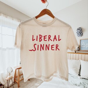 May include: Cream-colored, short-sleeved t-shirt with the words "LIBERAL SINNER" in red, with a red snake graphic. The shirt is hanging on a wooden hanger.