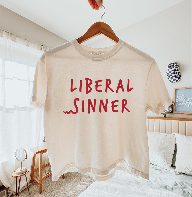 May include: Cream-colored, short-sleeved t-shirt with the words "LIBERAL SINNER" in red, with a red snake graphic. The shirt is hanging on a wooden hanger.