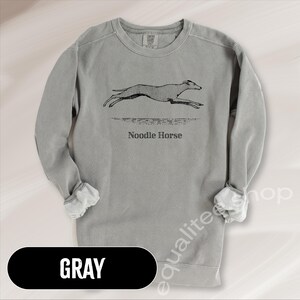 Pode incluir: Camisola cinzenta com um gr&aacute;fico preto de um c&atilde;o a correr e as palavras "Noodle Horse" impressas por baixo. A palavra "GRAY" est&aacute; impressa em branco num oval preto na parte inferior da imagem.