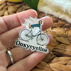 May include: A clear acrylic pin featuring a dachshund riding a bicycle. The dachshund wears a blue and green striped sweater. The pin has the word "Doxycycline" in a white font.