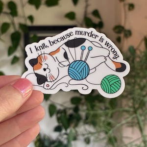 May include: A sticker featuring a cartoon cat with orange, black, and white markings, knitting with blue yarn. The text reads, "I knit, because murder is wrong." A green yarn ball is also present.