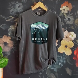 May include: A dark gray t-shirt with a graphic of a mountain range and a wolf howling at the moon. The graphic is in a blue and green color scheme and is surrounded by a dark blue hexagon with the text "DENALI FOREVER" underneath.