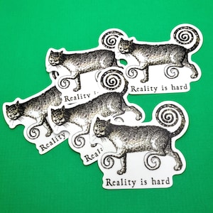 May include: A collection of cat stickers with a vintage illustration style. The cats are depicted in shades of gray and black, with a whimsical, curly tail design. Each sticker has the text "Reality is hard".
