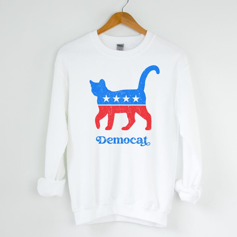 K&ouml;nnte beinhalten: Wei&szlig;er Pullover mit einer blauen und roten Katzensilhouette, die ein Design aus Sternen und Streifen tr&auml;gt. Der Text "Democat" ist unter der Katze aufgedruckt.
