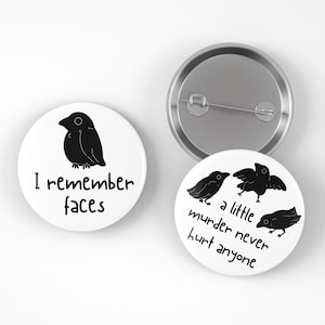 May include: White round button pins with black crow illustrations and text. One pin reads "I remember faces" and another says "a little murder never hurt anyone". A silver pin back is visible.