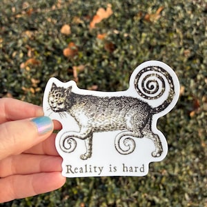 May include: A white sticker featuring a detailed illustration of a cat with a swirled tail and paws. The cat is in black and white. The text "Reality is hard" is printed below the cat.