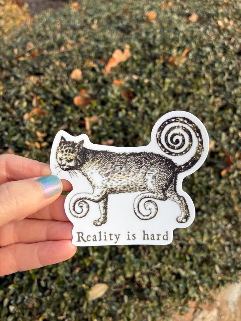 May include: A white sticker featuring a detailed illustration of a cat with a swirled tail and paws. The cat is in black and white. The text "Reality is hard" is printed below the cat.