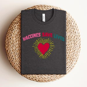 May include: A dark grey t-shirt with the text "Vaccines Save Lives" in pink, yellow, and teal lettering. A red heart with a yellow sunburst is centred on the shirt.