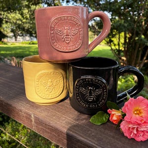 May include: Three ceramic mugs in dusty rose, mustard yellow, and black. Each mug features a circular embossed medallion with a bee illustration and the text BE BRAVE, BE LOUD, LOOK OUT FOR OTHERS. The mugs have a smooth finish and a side handle.