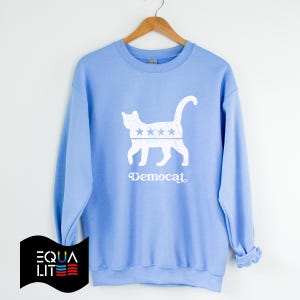 K&ouml;nnte beinhalten: Hellblauer Pullover mit einer wei&szlig;en Silhouette einer Katze mit Sternen auf dem K&ouml;rper und dem Wort "Democat" darunter.