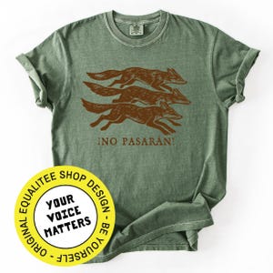 May include: Sage green t-shirt featuring a brown illustration of three running foxes. The Spanish phrase "¡NO PASARÁN!" is printed below the foxes. A round yellow sticker with the text "YOUR VOICE MATTERS" is in the lower left corner.