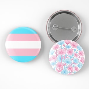 May include: Three round pinback buttons. One button features the transgender pride flag in light blue, white, and pink. Another button has a floral design in pink and blue. The third button is the back of a pin.