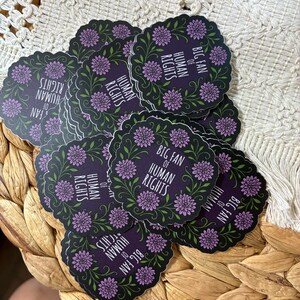 May include: A stack of round stickers with a dark purple background. Each sticker features a floral wreath design with purple flowers and green leaves. The text "BIG FAN OF HUMAN RIGHTS" is printed in white.