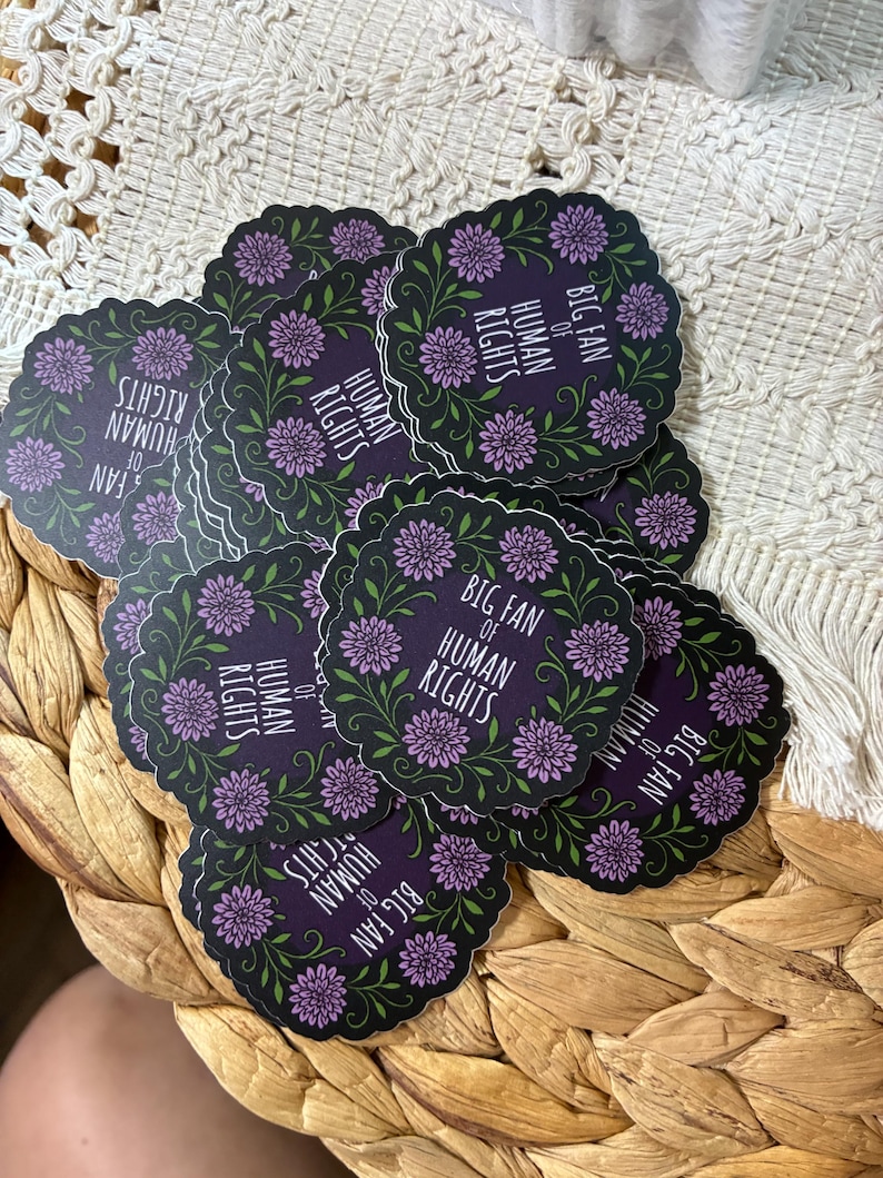 May include: A stack of round stickers with a dark purple background. Each sticker features a floral wreath design with purple flowers and green leaves. The text "BIG FAN OF HUMAN RIGHTS" is printed in white.