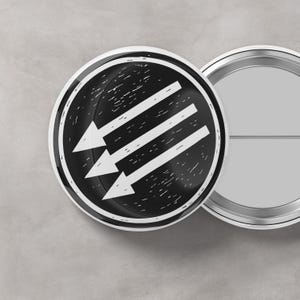 Puede incluir: Dos insignias redondas en blanco y negro. La insignia delantera tiene un fondo negro con tres flechas blancas que apuntan hacia abajo y hacia la izquierda. La segunda insignia es plateada y tiene un alfiler en la parte posterior.