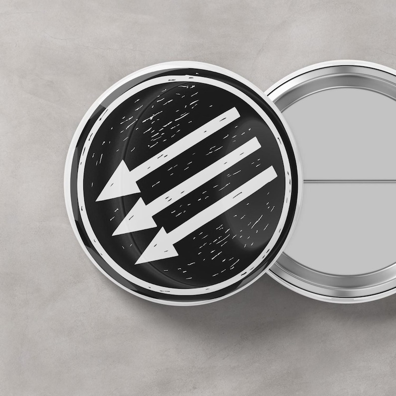 Puede incluir: Dos insignias redondas en blanco y negro. La insignia delantera tiene un fondo negro con tres flechas blancas que apuntan hacia abajo y hacia la izquierda. La segunda insignia es plateada y tiene un alfiler en la parte posterior.