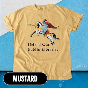 May include: A mustard yellow t-shirt featuring a graphic of a knight on horseback holding a spear and shield. The text "Defend Our Public Libraries" is printed below the graphic.