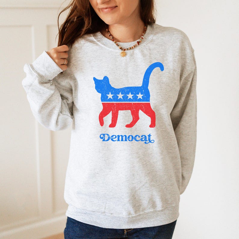 K&ouml;nnte beinhalten: Ein graues Sweatshirt mit einer roten, wei&szlig;en und blauen Katzensilhouette mit Sternen auf dem R&uuml;cken. Die Katze tr&auml;gt ein blaues Halsband mit einem Stern. Der Text "Democat" ist unter der Katze aufgedruckt.