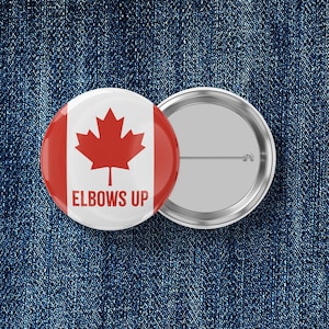 May include: A round button with a red maple leaf on a white background, with the text "ELBOWS UP" in red. The button has a silver metal backing. The background is a blue denim texture.