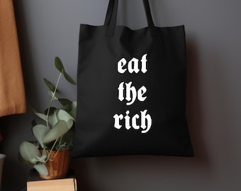 以下が含まれることがあります： 黒いトートバッグ。白いゴシック体で「eat the rich」の文字がプリントされています。バッグは灰色の壁に掛けられています。背景には木製の表面と鉢植えの小さな植物が見えます。