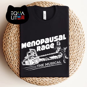 May include: Black t-shirt with a white graphic of a woman sitting on a chainsaw with the text "Menopausal Rage - The Musical".