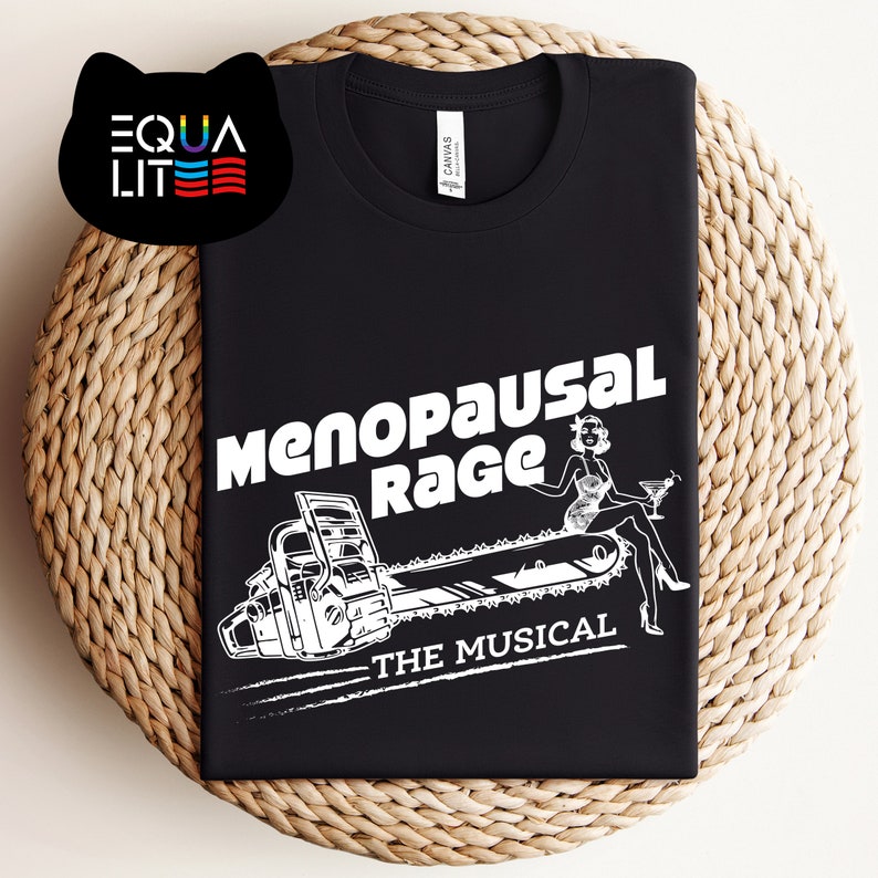 May include: Black t-shirt with a white graphic of a woman sitting on a chainsaw with the text "Menopausal Rage - The Musical".