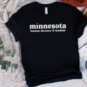 May include: Black t-shirt with white text that reads "minnesota human decency & hotdish" with a red heart above the word "minnesota".