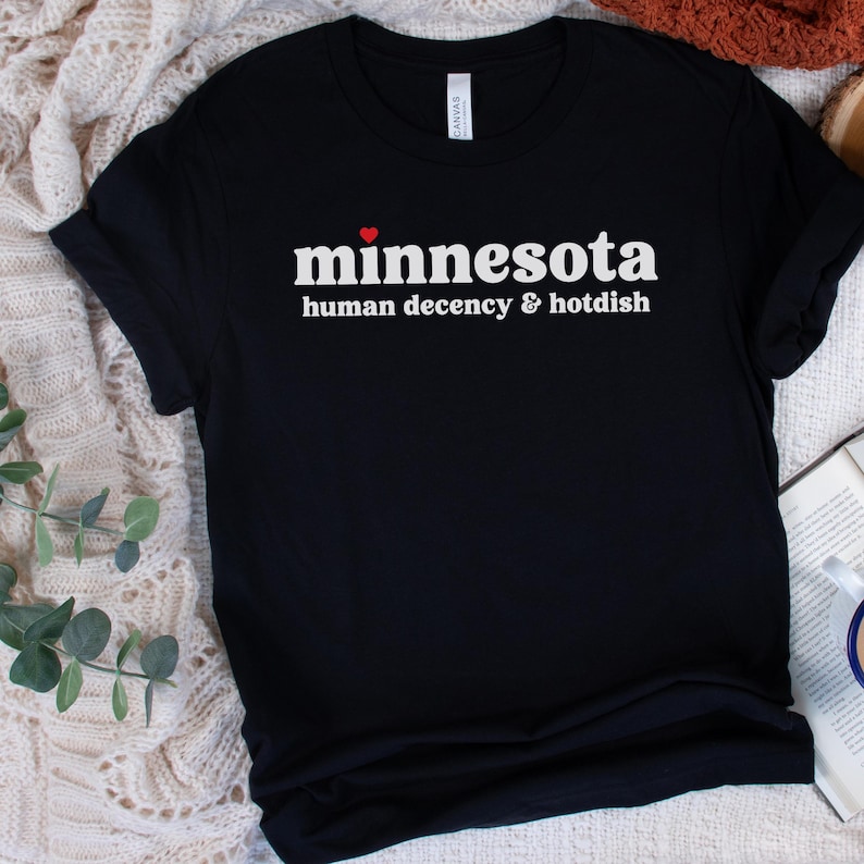 May include: Black t-shirt with white text that reads "minnesota human decency & hotdish" with a red heart above the word "minnesota".
